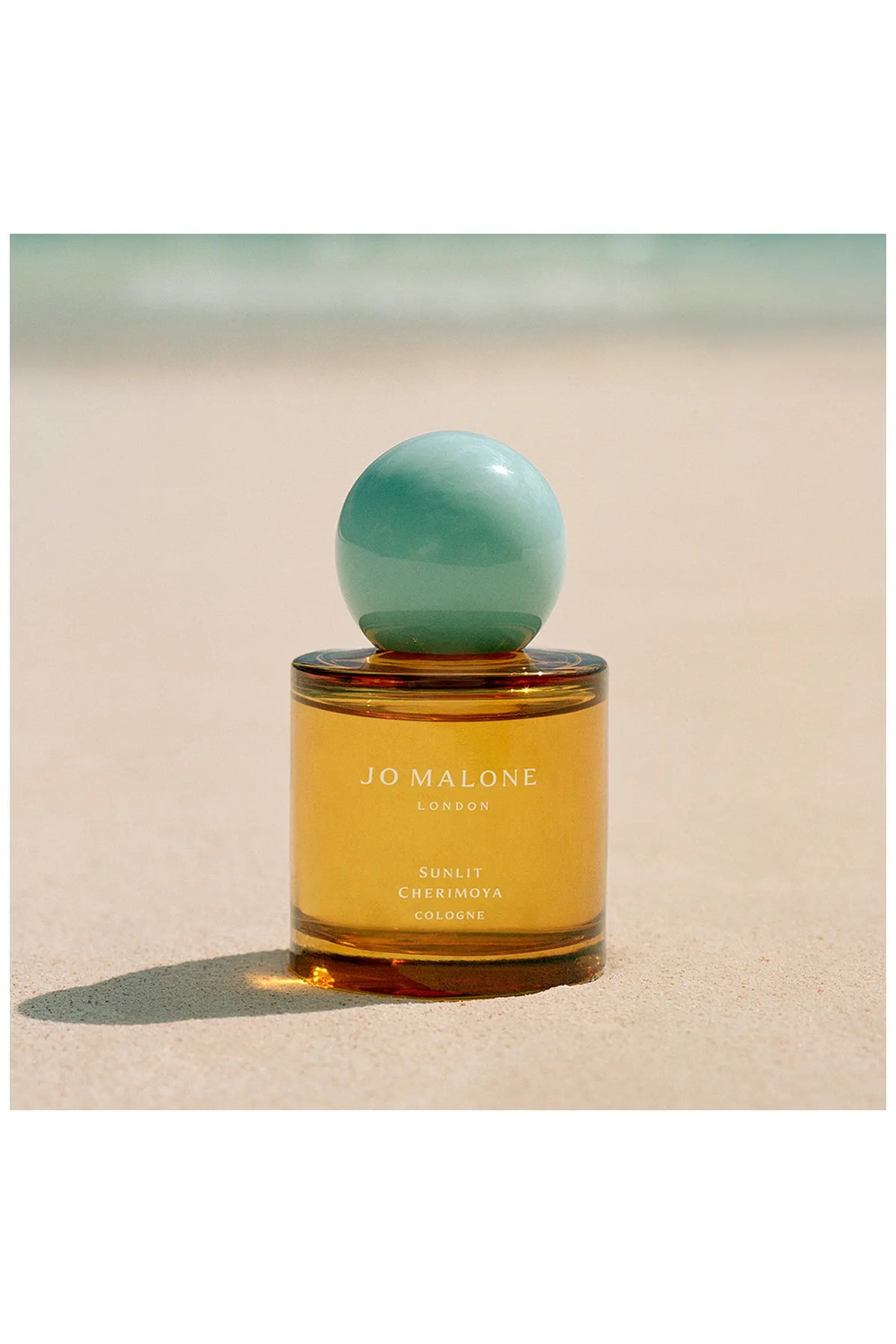 Jo Malone Sunlit Cherimoya Cologne im orangefarbenen Flakon auf hellem Sand in der Sonne.